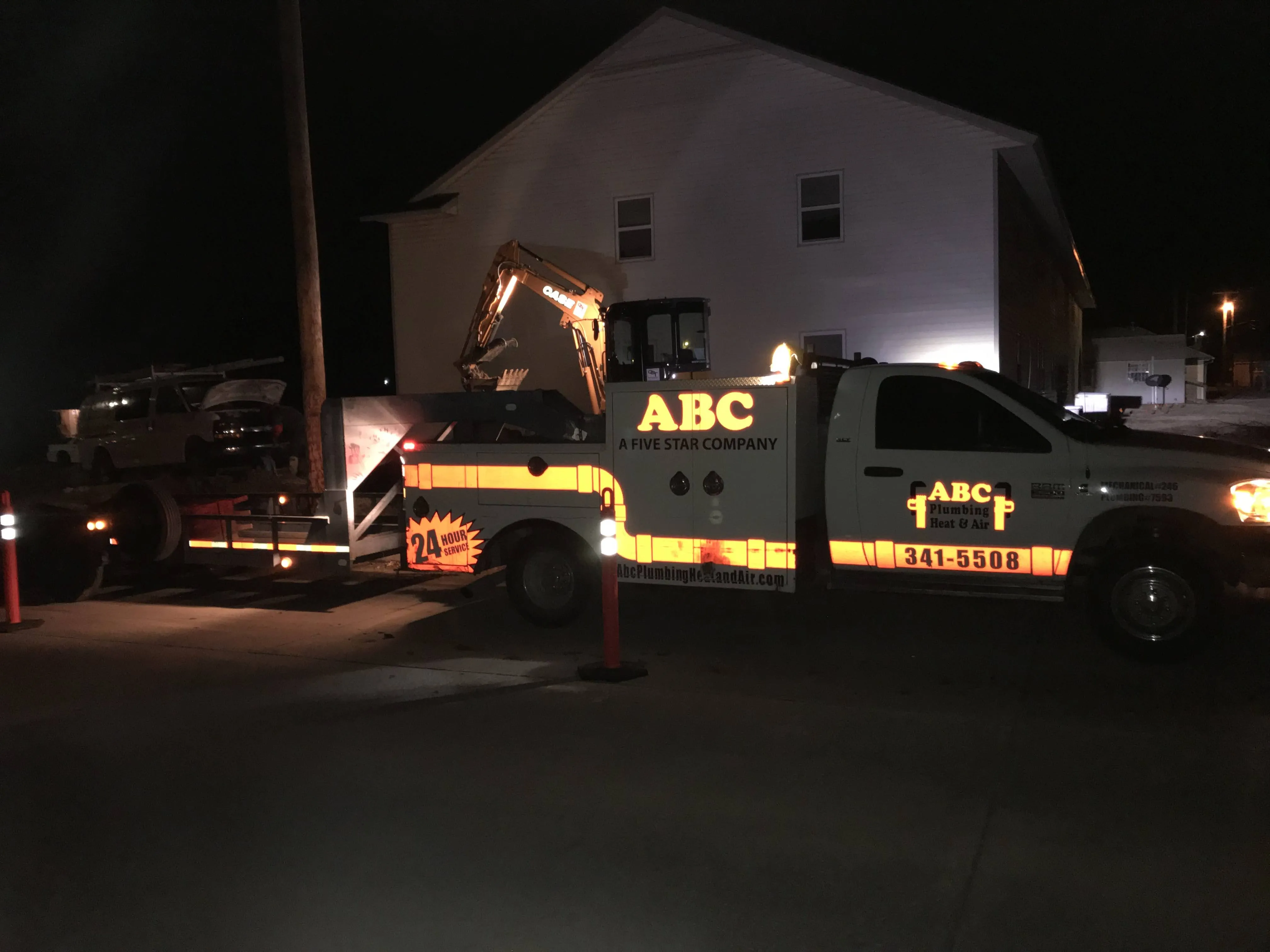 ABC Plumbing's branded truck and excavator on a trailer at a job site at night, with the 24-hour service logo and phone number lit up