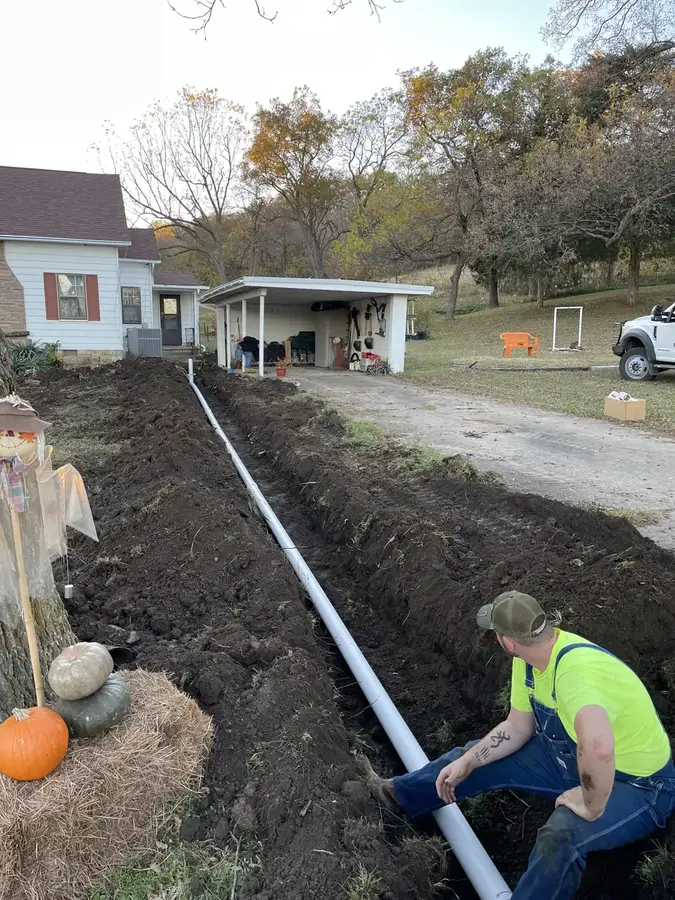 A crew member kneeling in a long trench beside a new white PVC sewer line running to a house