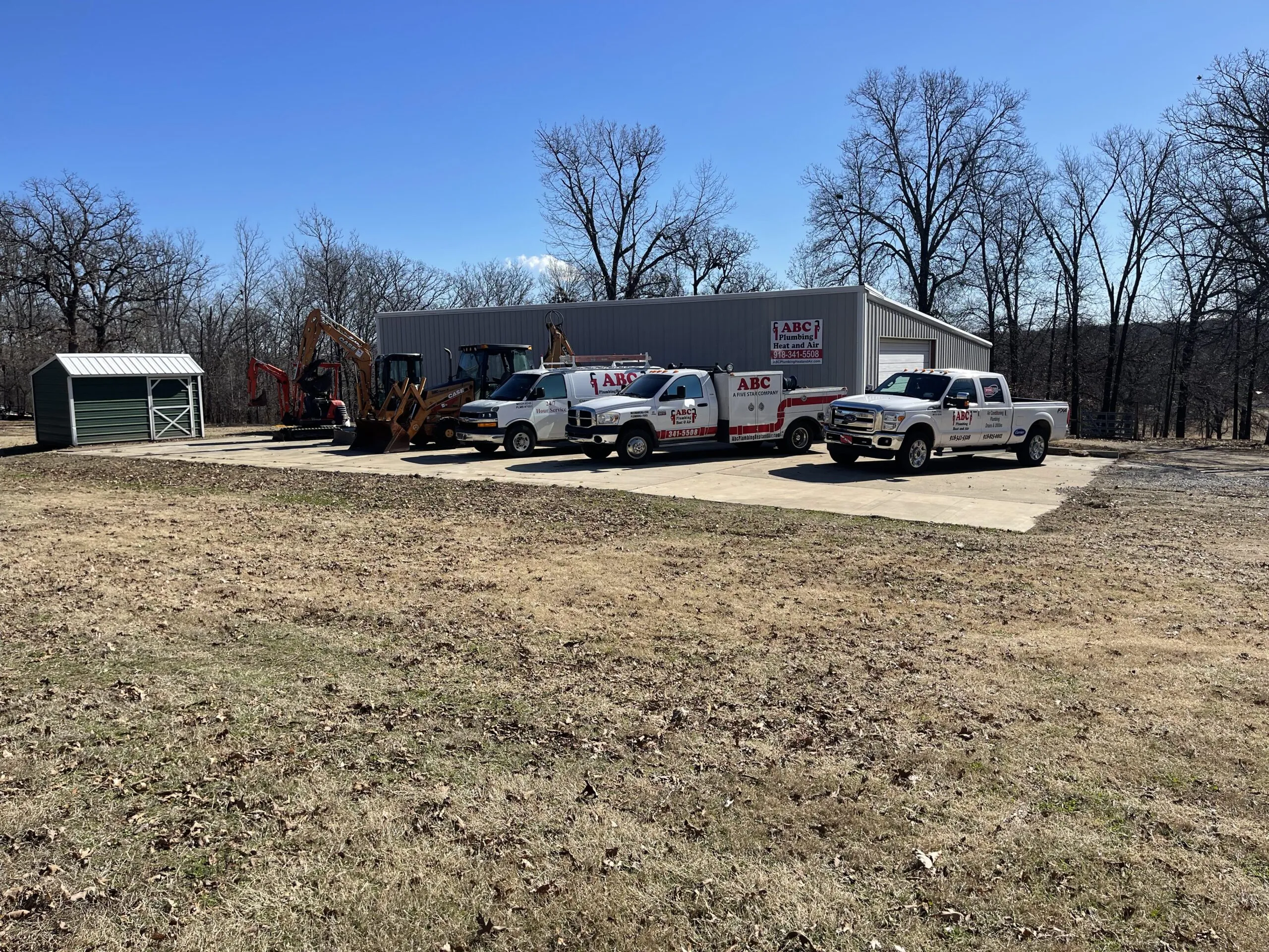ABC Plumbing Heat and Air's shop with the company sign, two branded trucks, the service van, and an excavator parked on the gravel lot